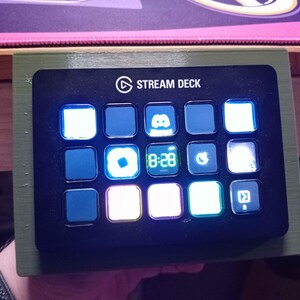 May include: A black Stream Deck with illuminated buttons, displaying various icons and the time 8:28. The device is mounted on a green wooden frame. The Stream Deck logo is visible at the top.