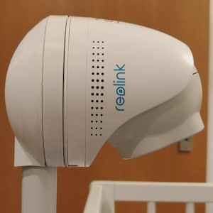 May include: A white Reolink security camera with a rounded design. The camera has a series of small holes on the side and the Reolink logo in blue. It is mounted on a white pole.