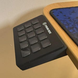 May include: Black Stream Deck with 15 illuminated buttons. The device is mounted on a wooden desk, with a blue and black patterned mousepad visible. The Stream Deck logo is printed on the side.