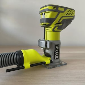 May include: A yellow and grey Ryobi One+ cordless router with a clear base. A black and grey flexible hose is attached. The router is on a light brown wooden surface.