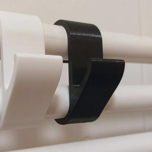 May include: Close-up of two hooks, one white and one black, hanging on a white pipe. The hooks are designed to hang items. The hooks are made of a smooth material and have a curved design.