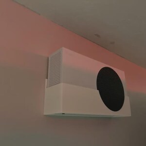 May include: A white rectangular electronic device with a large black circular vent is mounted on a light pink wall. The device has a perforated vent on the left side.
