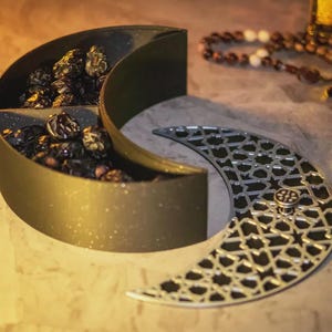 May include: A gold and silver crescent-shaped serving dish with a decorative lid, filled with dried fruit. The lid has an openwork design. A string of prayer beads is visible in the background. The dish is likely used for serving snacks.
