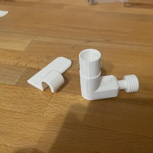 May include: White 3D-printed plastic components, including an L-shaped piece with a threaded end and a cylindrical opening, and a separate clip. The items are arranged on a wooden surface.