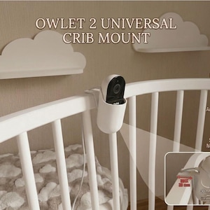 May include: A white crib with a baby monitor attached to the rail. The monitor is held in place by a white bracket. The text "OWLET 2 UNIVERSAL CRIB MOUNT" is displayed above the crib. Cloud-shaped shelves are also visible in the image.
