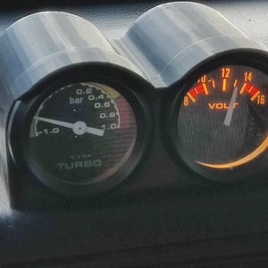 May include: A dual gauge instrument panel in a black housing. One gauge displays pressure in "bar", the other shows voltage. The gauges have white needles and orange backlighting. The text "TIM TURBO" is visible on the left gauge.