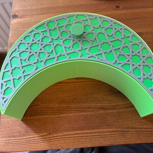 May include: A green, crescent-shaped decorative box with a gray geometric pattern. The box has a small, round knob on top. The box is made of a solid material and is on a wooden surface.