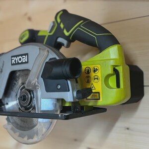 May include: A grey and lime green Ryobi circular saw with a black handle. The saw blade is visible, and the tool is mounted on a wooden surface. The saw has a black dust collection port.