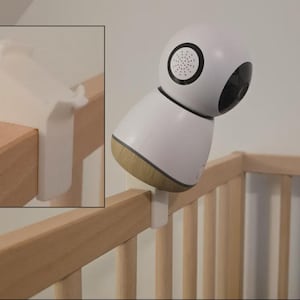 May include: A white baby monitor with a black and grey speaker and camera is attached to a wooden cot. The monitor has a rounded top and a wooden base. A white clip secures the monitor to the cot.