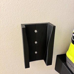 May include: Black plastic wall-mounted holder with three screws. The holder is designed to hold a power tool. The holder is mounted on a textured white wall.
