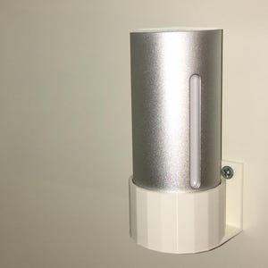 May include: A silver cylindrical device with a vertical illuminated strip, mounted on a white, geometric-shaped base. The device is attached to a wall with a screw. The design is modern and minimalist.
