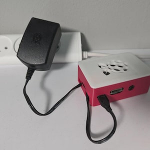 May include: A Raspberry Pi computer in a white and red case, connected to a black power adapter via a black cable. The power adapter is plugged into a white power strip.