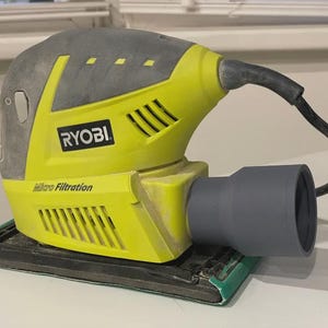 May include: A yellow and grey Ryobi Micro Filtration sander. The sander has a black power cable and a grey dust collection port. The base is black with a green edge. The Ryobi logo is visible.