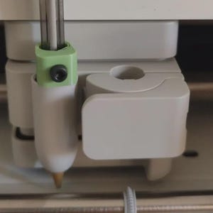 May include: A light grey plastic pen adapter for a cutting machine, featuring a pale green collar with a black metal screw. The adapter is mounted within the machine's tool housing, which includes a white plastic clamp mechanism for securing the pen.