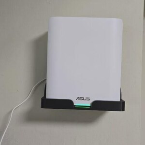 May include: A white ASUS router mounted on a wall. The router is rectangular with rounded corners and sits in a black holder. The ASUS logo is visible, and a green light is illuminated.