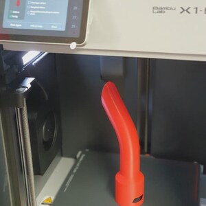 May include: A red, curved 3D-printed object is in progress inside a Bambu Lab X1-E 3D printer. The printer's screen shows a 'Completed' status. The object has a cylindrical base and a curved, elongated top.