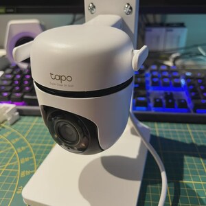 May include: White Tapo security camera with a black lens, mounted on a white stand. The camera is labelled "tapo Super Clear 3K SMP". The stand is attached to a white mounting plate with four screws.