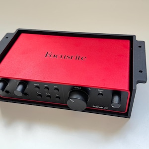 May include: A red Focusrite Scarlett 2i2 audio interface with black knobs and controls, set in a black frame. The Focusrite logo is visible on the top. The device has multiple input and output options for audio recording.