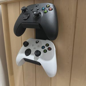 May include: Two Xbox controllers, one black and one white, mounted on a wooden surface. The controllers are held in place by a black bracket. The black controller is on top, and the white controller is below.