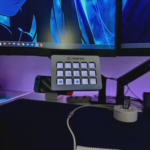 May include: A grey Stream Deck control panel with multiple customisable buttons, mounted below a computer monitor. The panel has a logo and various icons on the buttons. A Nintendo Switch and other tech accessories are visible in the background.