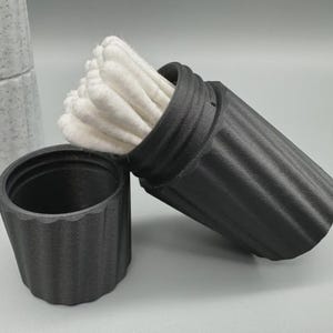 May include: A black, ribbed, cylindrical container with a lid, holding a bundle of white cotton buds. The lid is off, and the container is tilted, displaying the buds. A matching container is next to it. A stack of white, speckled containers is in the background.