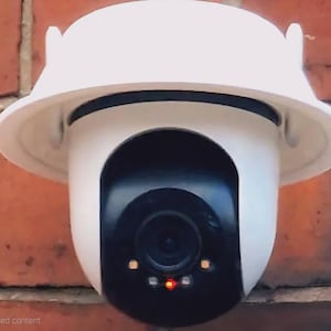 May include: A white and black security camera with a dome-shaped top and a black lens. The camera is mounted on a red brick wall. The camera has a red light and two orange lights.