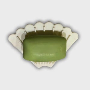 May include: A white, shell-shaped soap dish holds a rectangular, green bar of soap. The dish has a scalloped edge and is designed to allow water drainage. The soap is a solid, opaque green colour.