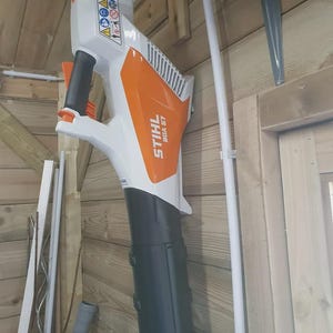 May include: A white and orange STIHL BGA 57 leaf blower. The blower has a black nozzle and handle. Safety symbols are visible on the top of the blower. The blower is mounted on a wooden wall.