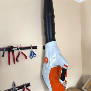 May include: A white and orange STIHL leaf blower is mounted on a beige wall. The blower has a black nozzle and handle. Various tools, including pliers and scissors, are mounted on the wall to the left.