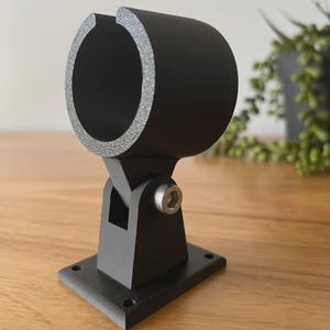 May include: A black, adjustable bracket with a circular opening. The bracket is mounted on a square base and features a pivoting joint secured by a silver bolt. The object is made of a matte black material and sits on a wooden surface.