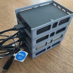 May include: A stack of four grey 3D-printed external hard drive enclosures. Each enclosure holds a black hard drive with a textured top. USB cables are connected to the drives. A blue and white sticker is attached to one of the cables.