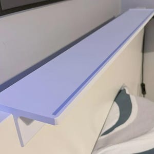 May include: A pale purple shelf fixed to a wall, running horizontally. It's supported by a white structure. A black lamp and a pillow with a blue and grey pattern are in the background. The shelf is designed to fit over a bed, offering storage.