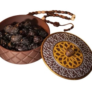 May include: A brown decorative container with a patterned lid, filled with dark dates. A beaded strand is draped over the container. The lid features a gold and silver design.