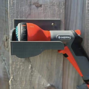 May include: A GARDENA brand garden hose nozzle and holder, in grey and orange, mounted on a wooden fence. The nozzle is red and black with a blue spray head. The holder is metal.