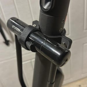 May include: A close-up shot of a black electric scooter's handlebar, featuring a black accessory secured with a velcro strap. The accessory has the word "KEEPER" printed on it. The scooter's stem and other parts are also black.