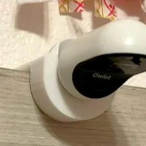 May include: A white and black Owlet baby monitor, mounted on a wall. The monitor has a rounded top and a black lens. The word "Owlet" is printed on the front of the device. The monitor is attached to a white circular base.
