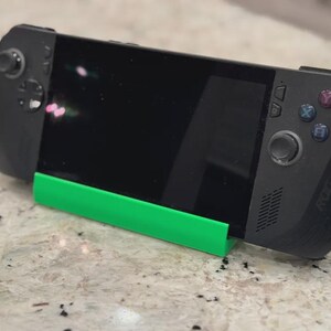 May include: A black handheld gaming device is supported by a bright green stand. The device has a black screen and buttons on either side. The stand is rectangular and appears to be made of plastic.