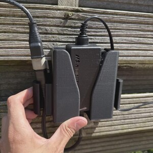 May include: A black electronic device with attached cables is mounted on a wooden fence. The device is held in place by a black holder. The device has the text "A V" printed on it.