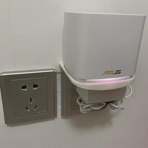 May include: A white ASUS router with a gold ASUS logo, mounted on a wall next to a silver electrical socket. The router has a curved design and a glowing light.