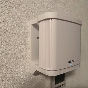 May include: A white ASUS network device mounted on a textured wall. The device has a rectangular shape with vertical lines on the front and the ASUS logo. A small blue light is on.