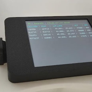 May include: A black electronic device with a screen displaying data. The device has a rectangular shape with rounded corners and a small cylindrical component attached to its side. The screen shows rows of text and numbers in a green font.