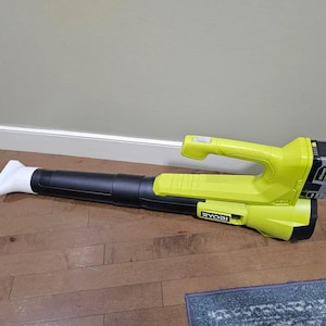 May include: A cordless leaf blower, mainly black with a bright yellow handle and details. The blower has a white nozzle and is lying on a wooden floor. The brand name "RYOBI" is visible.