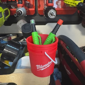 May include: A red Milwaukee bucket with a white handle contains two green and black tools with red and clear tips. The bucket is in a workshop, surrounded by other tools and equipment.