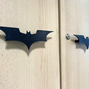 May include: Two black Batman logo cabinet pulls are mounted on a light wood cabinet. The bat-shaped handles are a dark matte colour, and one is near a silver keyhole. The cabinet doors have a natural wood grain.