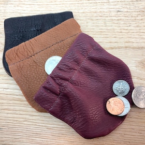 May include: Three small leather coin purses in black, brown, and burgundy. The burgundy purse holds several coins. The purses are made of textured leather and have a drawstring closure. The purses are displayed on a wooden surface.