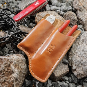 May include: A brown leather pouch with two slots for carrying tools. The pouch has the initials "FLS" embossed on the front. The pouch is sitting on a bed of small rocks.