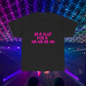 May include: Black t-shirt with the text "IM A SLUT FOR A HA HA HE HU" in bright pink. The shirt is a crew neck style and appears to be made of a soft material. The background is a concert stage with purple and blue lighting.