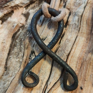 May include: A dark blue metal pendant in the shape of a stylized infinity symbol, hanging from a light brown leather cord. The pendant is on a wooden surface.