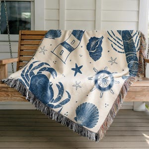 May include: A cream-colored throw blanket with a nautical theme, featuring navy blue illustrations of a crab, seashells, a lighthouse, starfish, and a ship's wheel. The blanket has a fringed edge and is draped over a wooden swing.