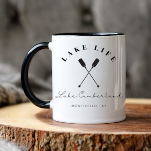 May include: White ceramic mug with a black handle and rim. The mug features the text "LAKE LIFE" above crossed oars, with "Lake Cumberland" and "Monticello - KY" below. The mug is on a wooden surface.